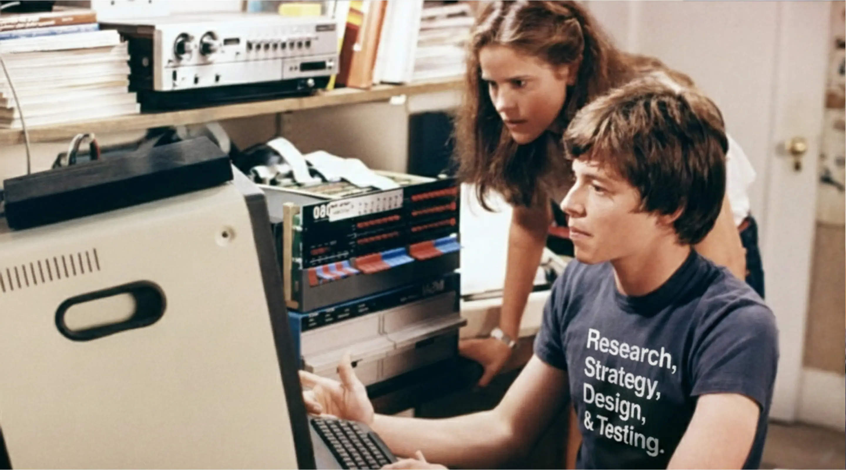 Still from WarGames, 1983 — a teenager at an early personal computer, t-shirt reading Research, Strategy, Design & Testing.
